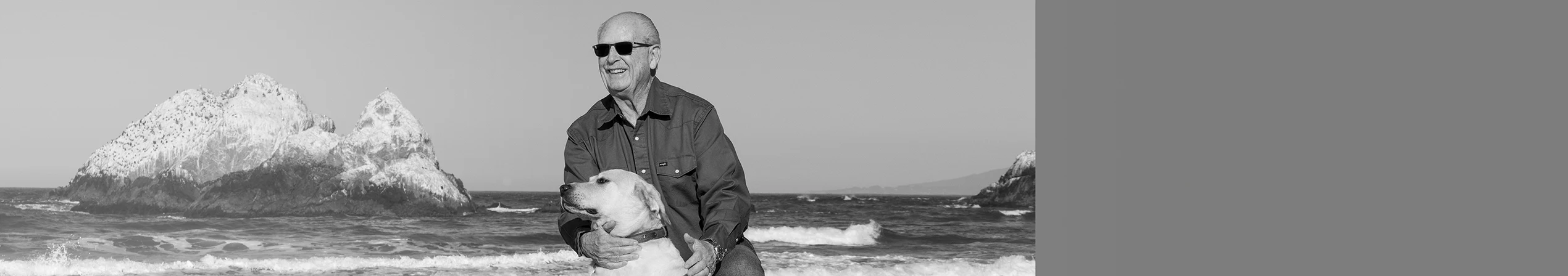 Man wearing sunglasses plays with his dog on a beach.