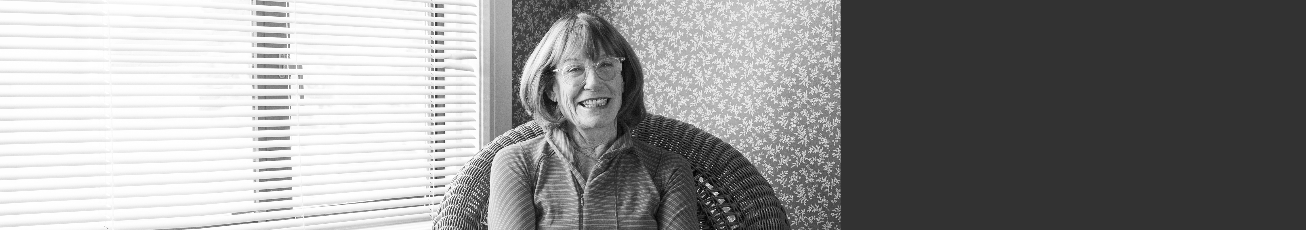 Bespectacled woman sits in rattan chair next to a window.