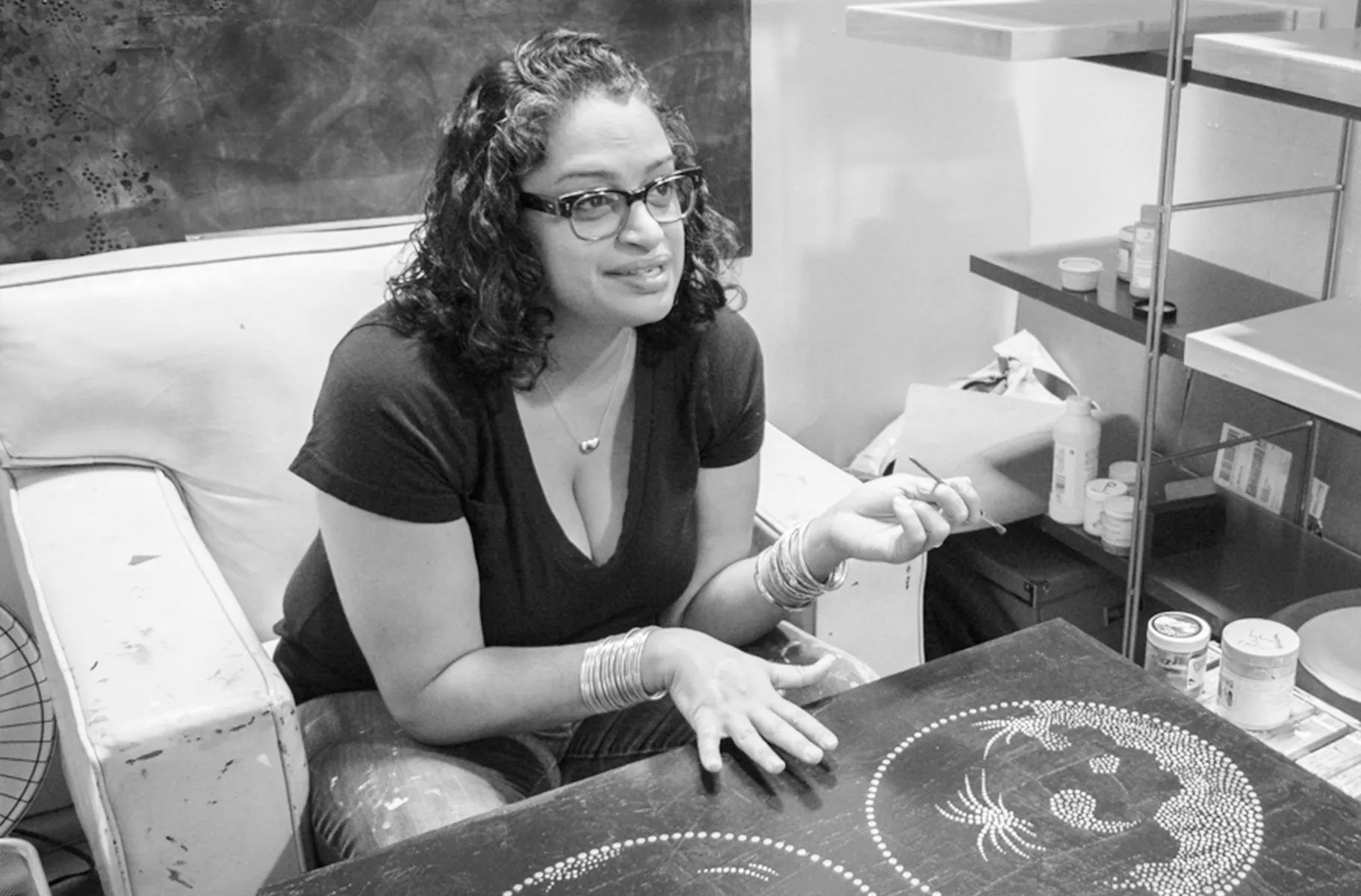 Woman sits in a large armchair with a painting on a the in front of her. She holds a paintbrush in one hand and is speaking excitedly.