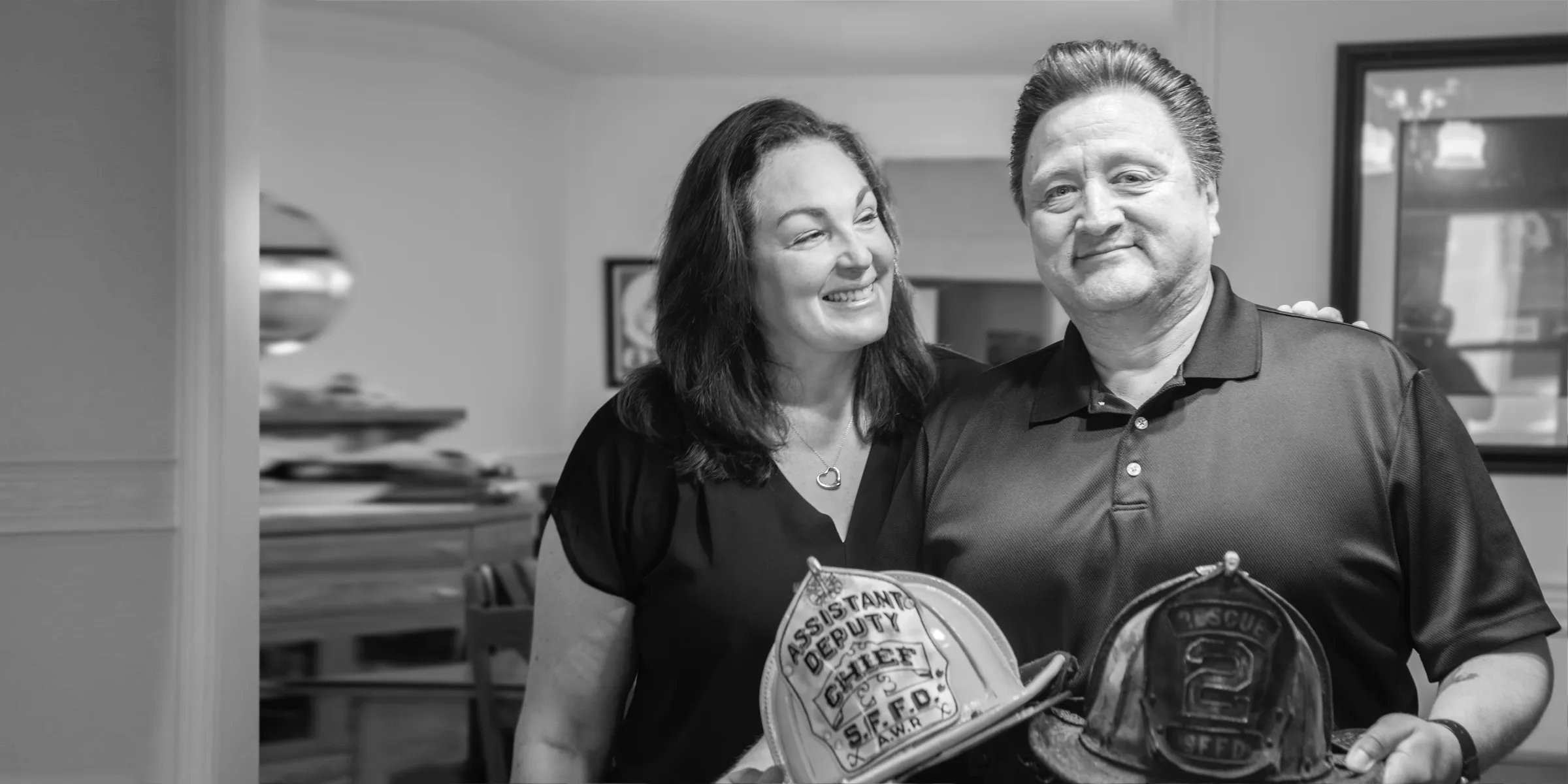 Adult man holds two fire helmets, showing them off to the camera. Adult woman is touching his shoulder and looking at him affectionately