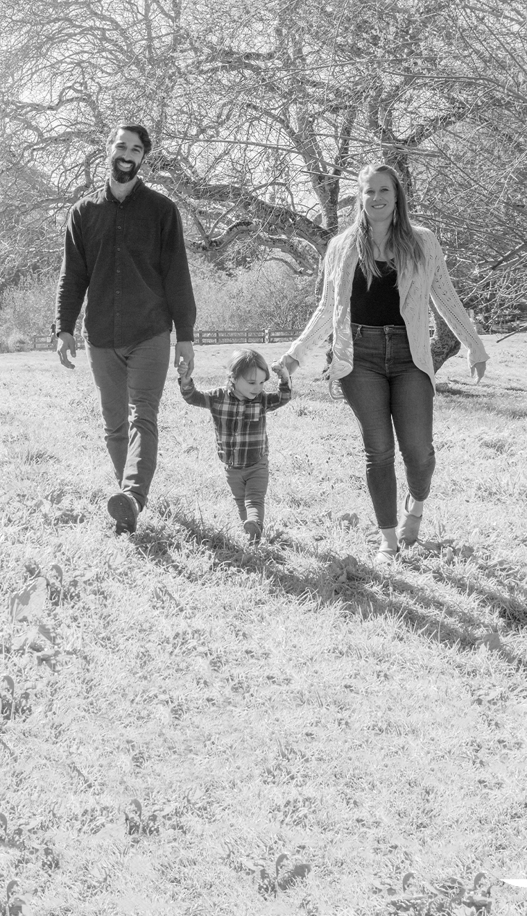 Parents stand on either side of their toddler, each holding one of the child's hands.